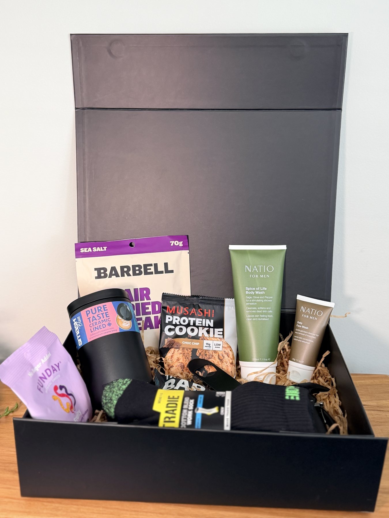 The Worksite Warrior Hamper packed open, showing air dried steak, tumbler, protein cookie, Natio wash, Tradie socks, a Funday sweet packet and multi-tool hammer
